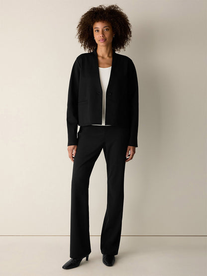 A woman stands against a plain background wearing the Eileen Fisher Extra-Fine Ponte Kick Pant in Black—crafted from the lightest ponte and featuring a subtle flare at the hem—paired with a black blazer, white top, and sleek black shoes.