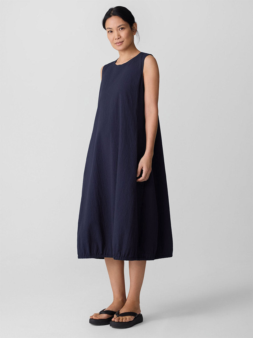 A woman stands against a plain background wearing the Eileen Fisher Organic Cotton Pucker Lantern Dress in Ink and black sandals.