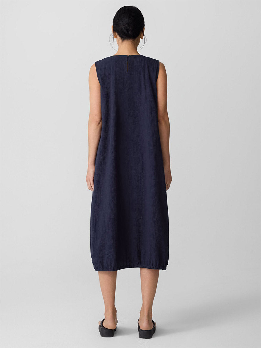 A person faces away, wearing the Eileen Fisher Organic Cotton Pucker Lantern Dress in Ink and black slip-on shoes against a plain light background.