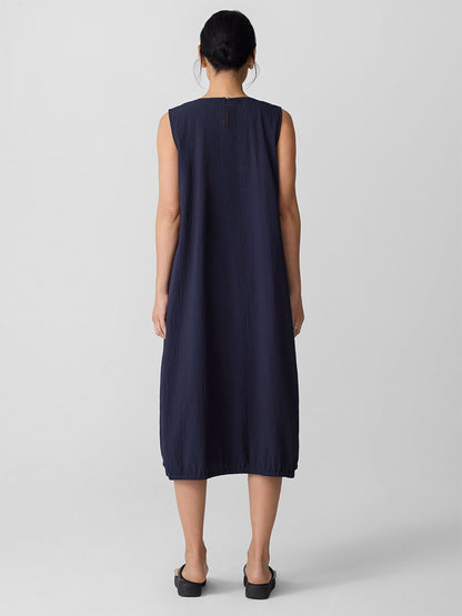 A person faces away, wearing the Eileen Fisher Organic Cotton Pucker Lantern Dress in Ink and black slip-on shoes against a plain light background.