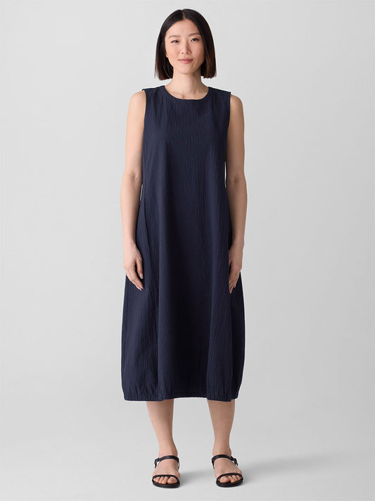 A woman faces forward, wearing the Eileen Fisher Organic Cotton Pucker Lantern Dress in Ink and black sandals, standing against a plain light grey background.