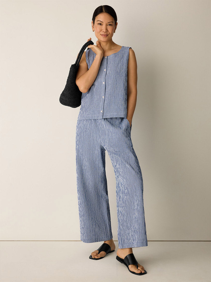 A woman stands against a plain background wearing a blue and white striped sleeveless top, Eileen Fisher Organic Cotton Pucker Check Wide-Leg Pant in Light Dive, black sandals, and holding a black woven bag.