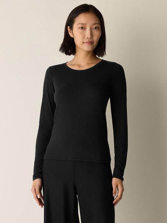 A woman with short dark hair wears the Eileen Fisher Sheer Cotton Blend Knit Round Neck Tee in black, paired with dark pants, standing against a plain light-colored background.