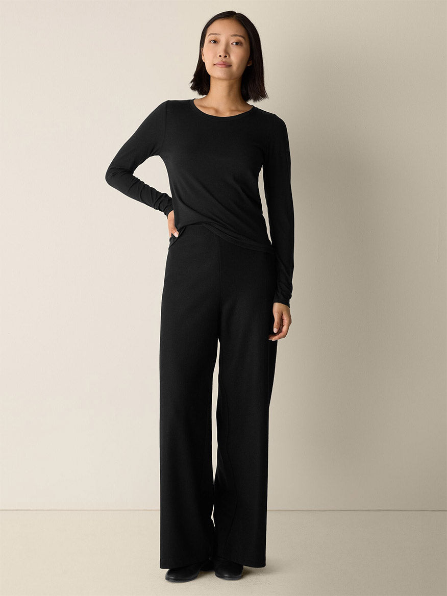 A woman stands against a plain light-colored wall, wearing the Eileen Fisher Sheer Cotton Blend Knit Round Neck Tee in Black with wide-leg black pants and black shoes.