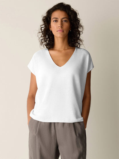 A woman with curly hair wears the Eileen Fisher Organic Linen Cotton V-Neck Top in White and gray pants, standing with hands in pockets against a plain light background.