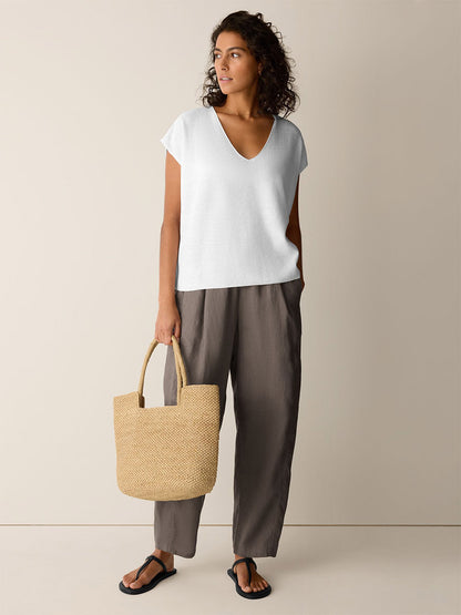 A woman stands indoors wearing the Eileen Fisher Organic Linen Cotton V-Neck Top in white, paired with loose brown pants, black sandals, and holding a woven straw bag.