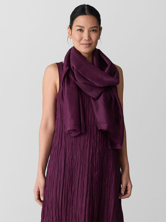 A woman wearing a sleeveless textured purple dress and the Eileen Fisher Washed Transitional Silk Scarf in Blackberry stands against a plain light background.