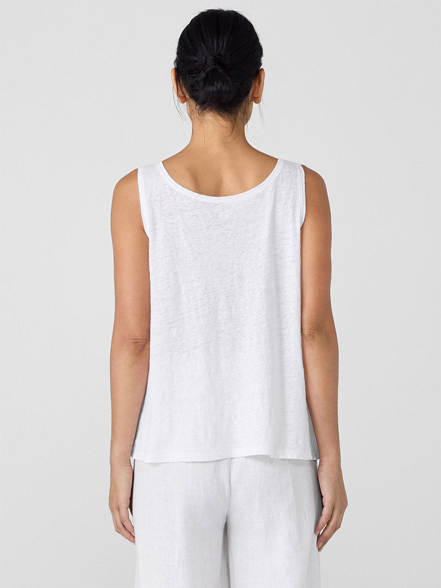 A woman with dark hair in a bun is shown from the back, wearing an Eileen Fisher Organic Linen Jersey Scoop Neck Tank in White and white pants against a plain light background.