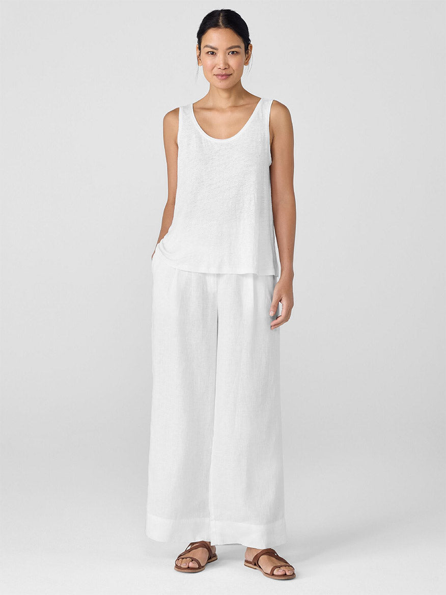 A woman faces forward wearing an Eileen Fisher Organic Linen Jersey Scoop Neck Tank in White, wide-leg white pants, and brown sandals against a plain light background.