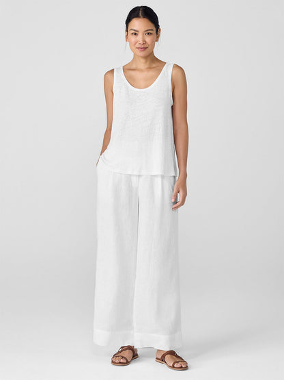 A woman faces forward wearing an Eileen Fisher Organic Linen Jersey Scoop Neck Tank in White, wide-leg white pants, and brown sandals against a plain light background.