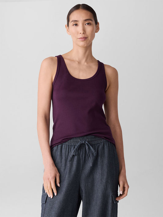 A woman stands against a plain background, wearing the Eileen Fisher Organic Cotton Stretch Rib Scoop Neck Tank in Blackberry and loose gray drawstring pants.
