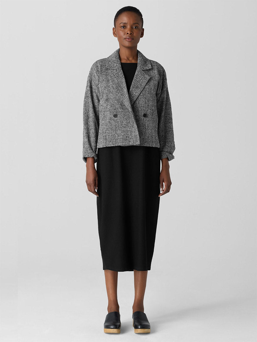 Set against a plain background, the individual looks stylish in a chic black dress paired with the elegant Eileen Fisher Organic Cotton Tweed Short Blazer in Black/White and black shoes. This sophisticated outfit blends fashion with conscious living and is certified Fair Trade.