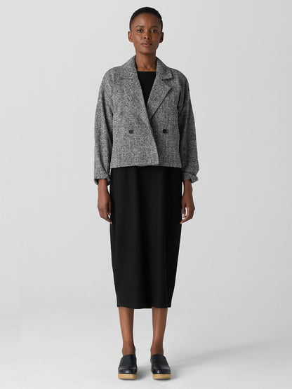 Set against a plain background, the individual looks stylish in a chic black dress paired with the elegant Eileen Fisher Organic Cotton Tweed Short Blazer in Black/White and black shoes. This sophisticated outfit blends fashion with conscious living and is certified Fair Trade.