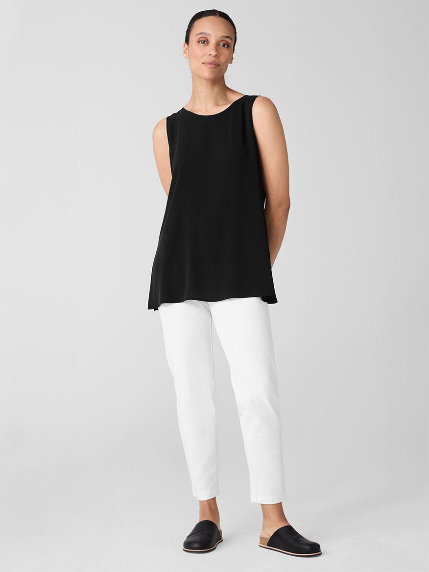 A person stands gracefully against a plain backdrop, wearing a sleeveless black top paired with chic Eileen Fisher Washable Stretch Crepe Pant in White and sleek black slip-on shoes, exuding an effortlessly stylish presence.