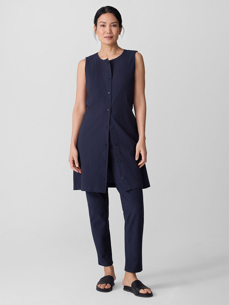A woman stands facing forward, wearing a sleeveless navy button-up tunic, Eileen Fisher Washable Stretch Crepe Pant in Ink, and black slide sandals against a plain light gray background.