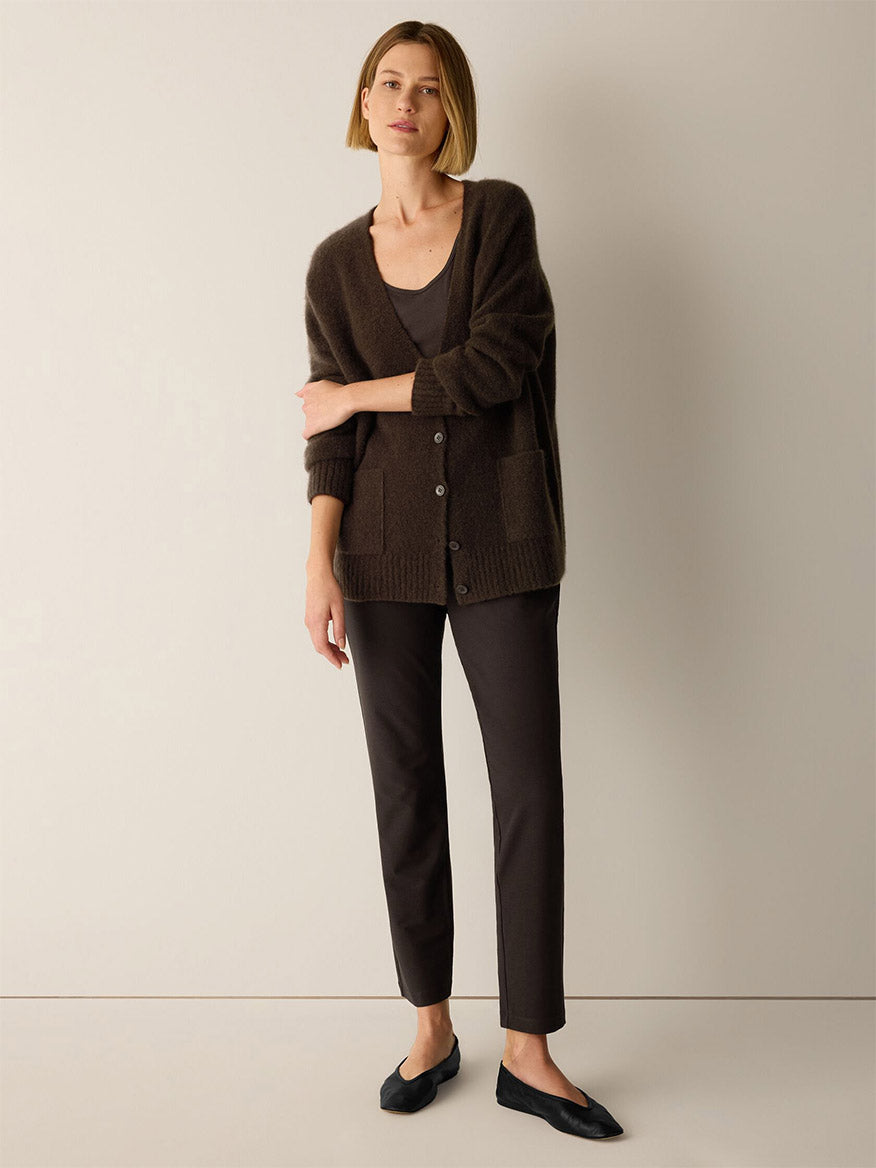 A woman wears a dark brown button-up cardigan over a grey scoop-neck top, paired with Eileen Fisher Washable Stretch Crepe Slim Pant in Coffee and black ballet flats.