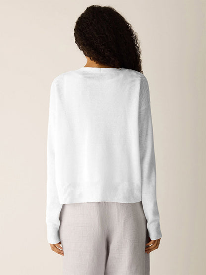A person with curly hair is shown from the back, wearing an Eileen Fisher Organic Linen Cotton Slouchy Cardigan in white with light-colored pants, standing against a plain background.