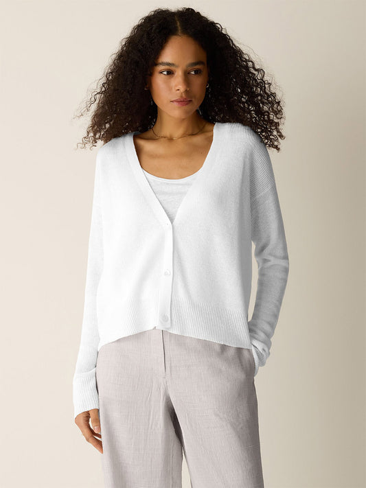 A woman with curly hair wears the Eileen Fisher Organic Linen Cotton Slouchy Cardigan in White over a light top and light gray pants, standing against a plain light background.