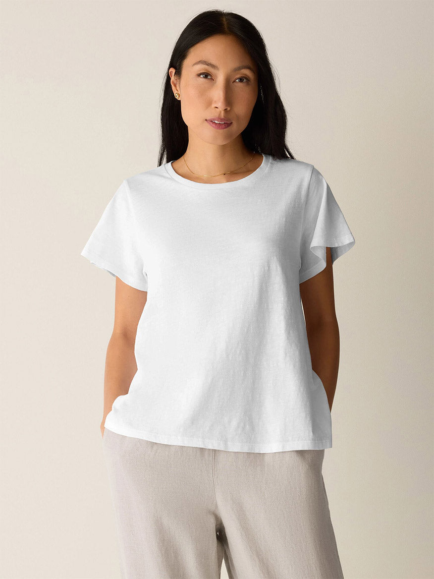 A woman stands against a plain background wearing the Eileen Fisher Organic Cotton Slubby Jersey Crew Neck Tee in White and light beige pants, hands in pockets.