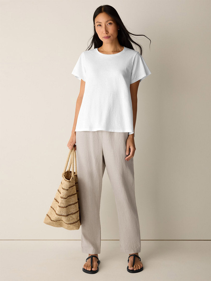 A woman stands against a plain background wearing the Eileen Fisher Organic Cotton Slubby Jersey Crew Neck Tee in White, paired with beige pants, black sandals, and holding a striped woven tote bag.