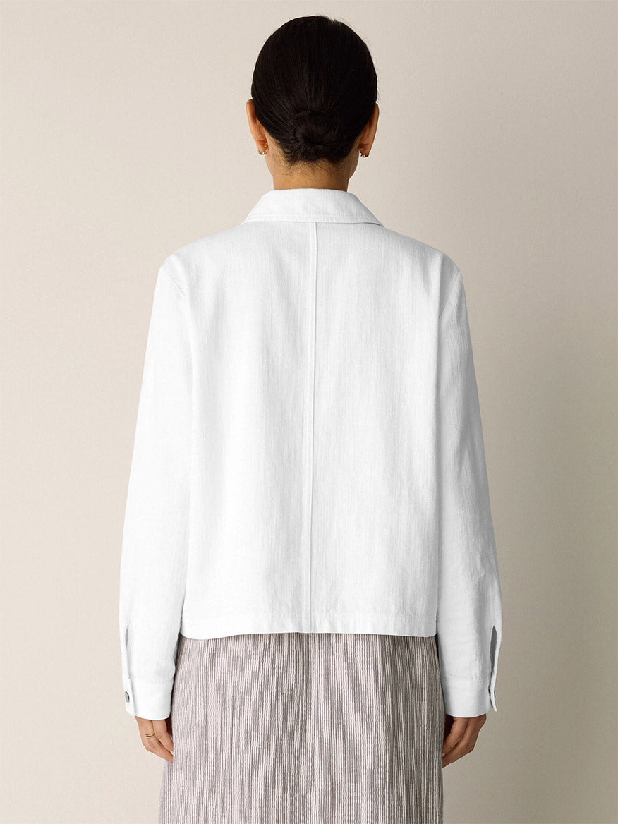A person stands facing away, wearing the Eileen Fisher Slubby Organic Cotton Linen Classic Collar Jacket in white, styled with a light pleated skirt against a plain light-colored background.