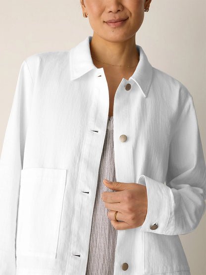 A person wearing the Eileen Fisher Slubby Organic Cotton Linen Classic Collar Jacket in White over a textured light top, with one hand touching the jacket near the buttons.