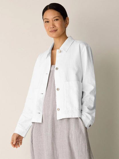 A woman stands against a plain background wearing the Eileen Fisher Slubby Organic Cotton Linen Classic Collar Jacket in white over a light gray dress, with her hand in her pocket and a neutral expression.