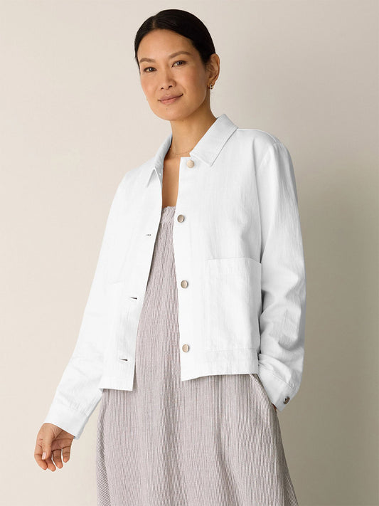 A woman stands against a plain background wearing the Eileen Fisher Slubby Organic Cotton Linen Classic Collar Jacket in white over a light gray dress, with her hand in her pocket and a neutral expression.