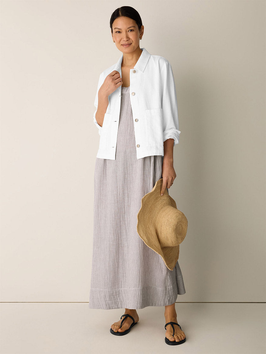A woman stands indoors, smiling at the camera, wearing a light gray organic cotton linen maxi dress, black sandals, and holding a wide-brimmed straw hat. She has on an Eileen Fisher Slubby Organic Cotton Linen Classic Collar Jacket in White.