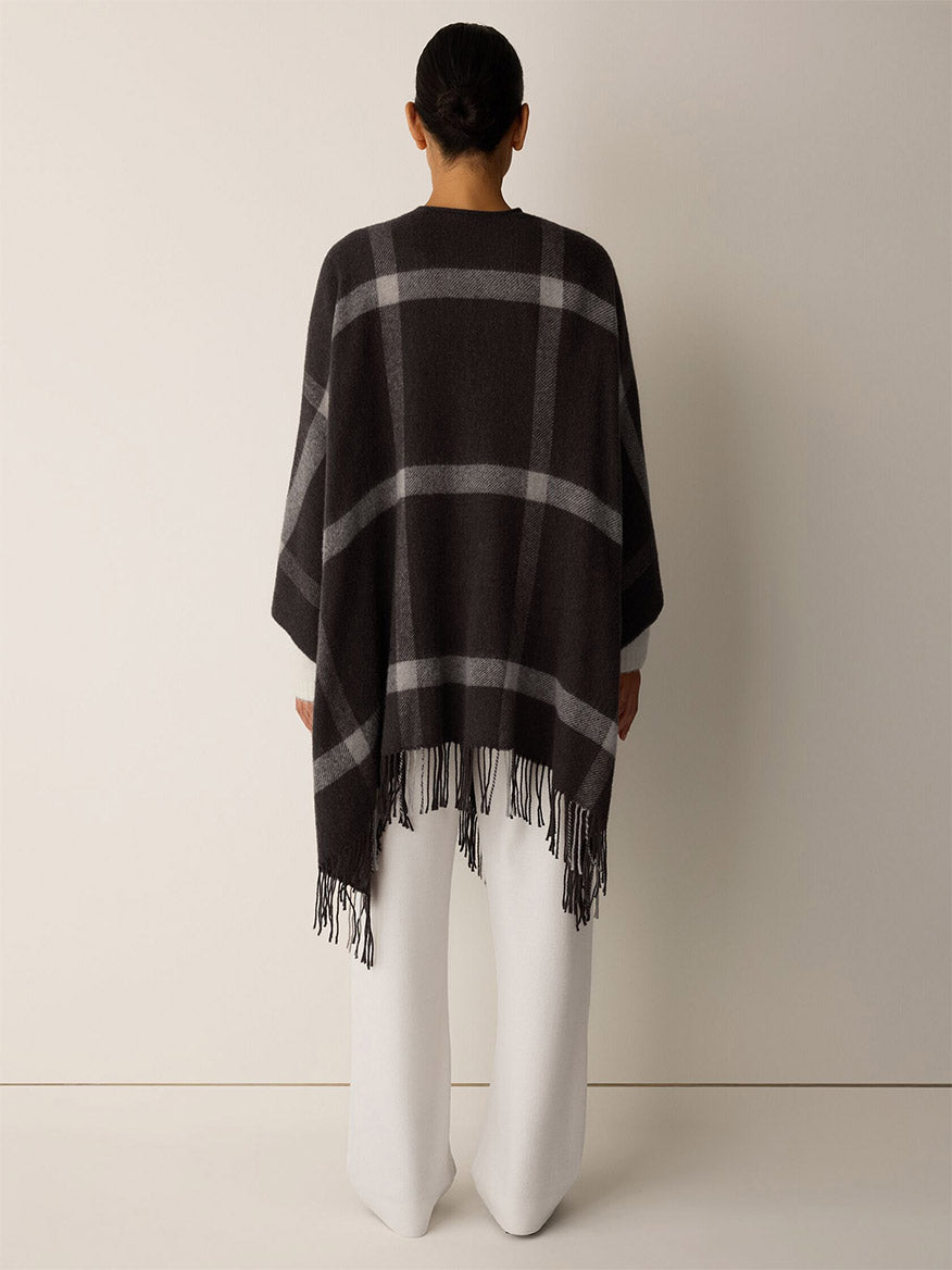 Back view of a person wearing an Eileen Fisher Soft Wool Plaid Serape in Coffee over white trousers, featuring a charcoal and gray graphic grid pattern with fringe trim.