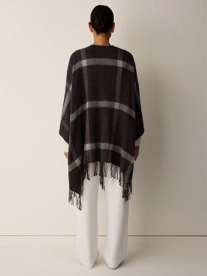 Back view of a person wearing an Eileen Fisher Soft Wool Plaid Serape in Coffee over white trousers, featuring a charcoal and gray graphic grid pattern with fringe trim.