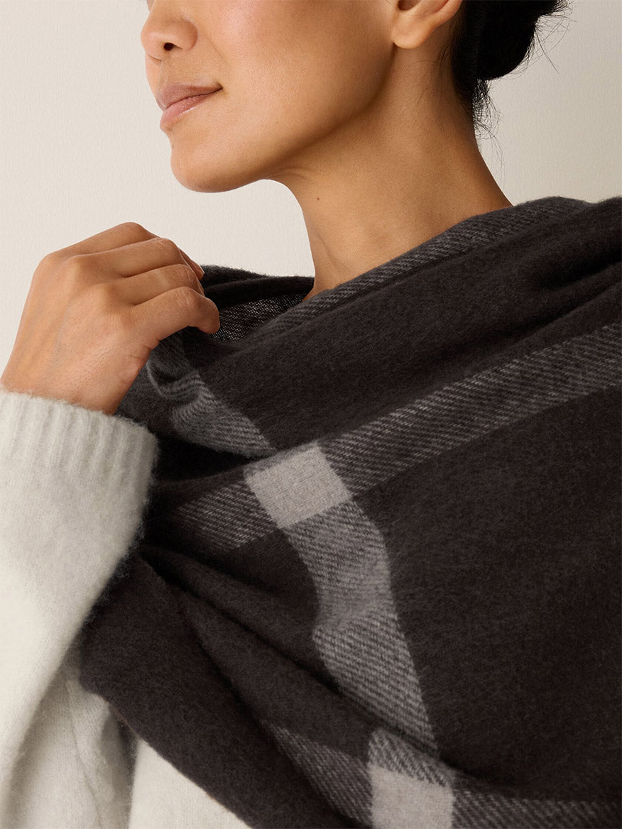 An Eileen Fisher Soft Wool Plaid Serape in Coffee featuring a dark brown and grey recycled wool graphic grid pattern draped over a person’s shoulders.