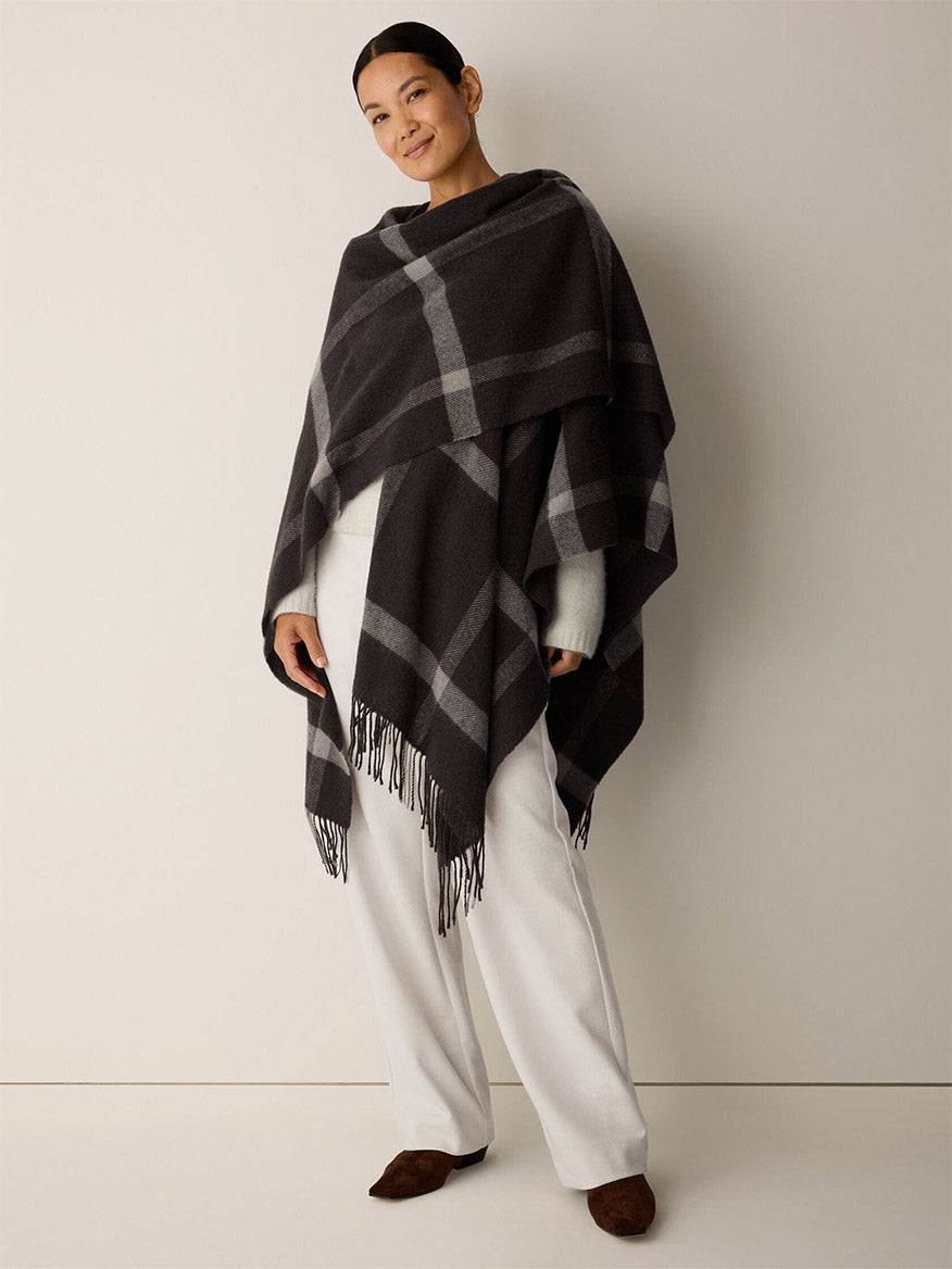 A person wearing an Eileen Fisher Soft Wool Plaid Serape in Coffee over a white sweater and trousers, featuring a dark brown graphic grid pattern and a fringe hem.