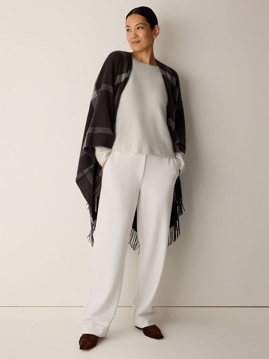 A person wears an Eileen Fisher Soft Wool Plaid Serape in Coffee featuring a graphic grid pattern over a light-colored sweater and white trousers.