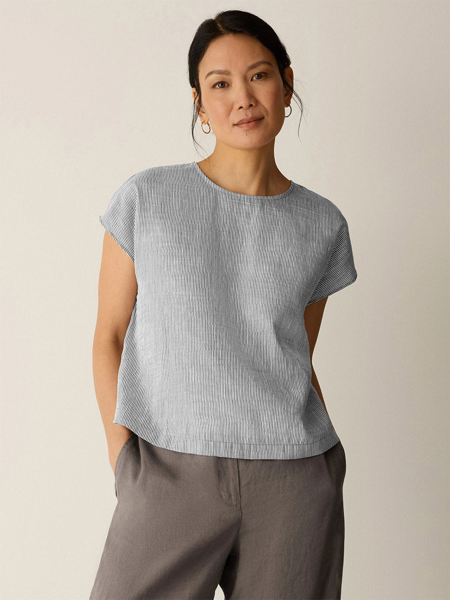 A woman stands against a plain background wearing the Eileen Fisher Striped Linen Sheen Square Top in Wheat, a boxy fit with subtle stripes, and loose grey pants, her hands in her pockets.