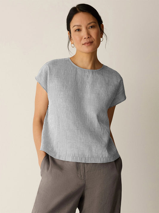 A woman stands against a plain background wearing the Eileen Fisher Striped Linen Sheen Square Top in Wheat, a boxy fit with subtle stripes, and loose grey pants, her hands in her pockets.