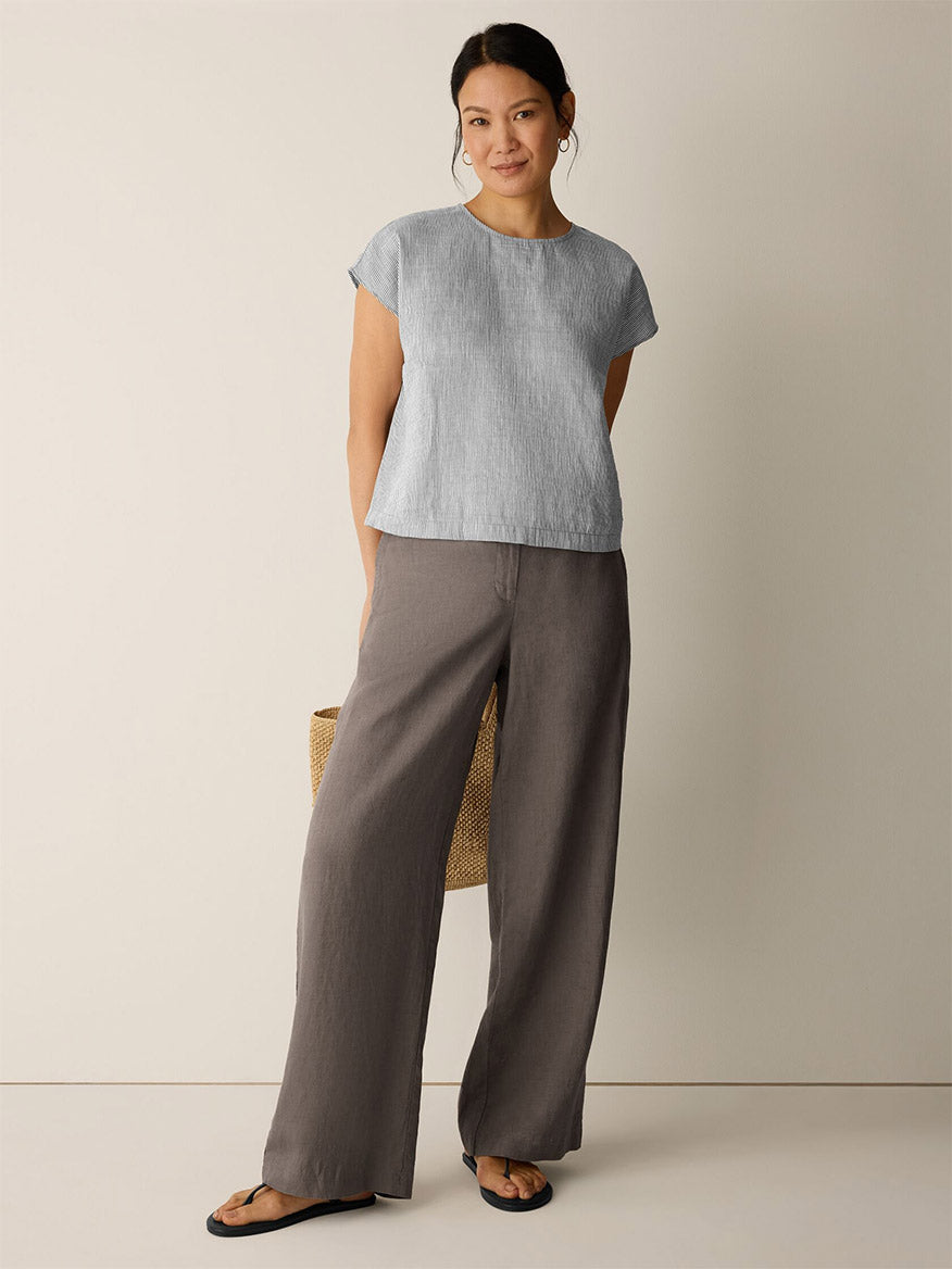 A woman stands against a plain wall wearing the Eileen Fisher Striped Linen Sheen Square Top in Wheat, wide-leg brown pants, black sandals, and holding a woven bag.