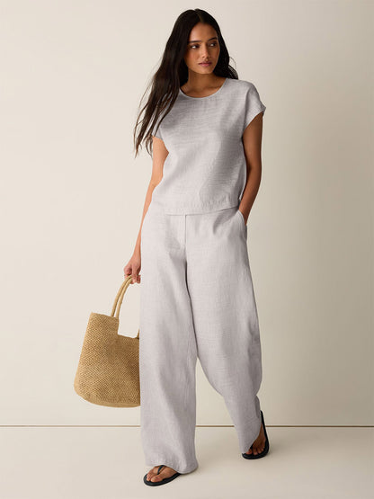 A woman stands against a plain backdrop wearing the Eileen Fisher Striped Linen Sheen Square Top in Wheat with a boxy fit, styled with wide-leg pants, a woven tote bag, and black sandals.