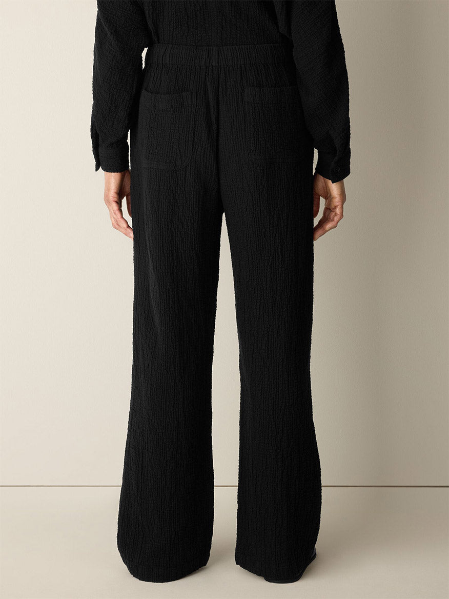 A person stands with their back to a beige wall, wearing the Eileen Fisher Organic Cotton Doubleweave Straight Pant in Black, featuring a relaxed fit and drawstring waist, paired with a matching black textured top.