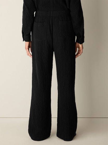 A person stands with their back to a beige wall, wearing the Eileen Fisher Organic Cotton Doubleweave Straight Pant in Black, featuring a relaxed fit and drawstring waist, paired with a matching black textured top.
