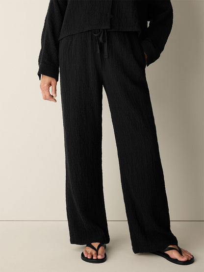 Wearing Eileen Fisher Organic Cotton Doubleweave Straight Pant in Black, featuring a relaxed straight fit and drawstring waist, styled with a matching long-sleeve top and black flip-flops, the person stands against a plain light background.