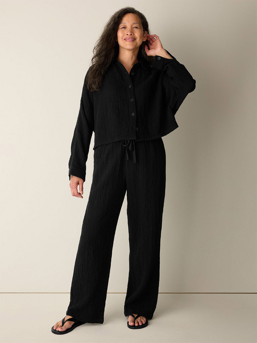 A woman stands against a plain wall, smiling with one hand touching her hair, wearing relaxed black sandals and the Eileen Fisher Organic Cotton Doubleweave Straight Pant in Black—a straight-leg style featuring a drawstring waist—paired with a loose black button-up top.
