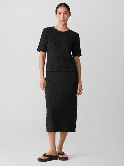 A woman stands against a plain background wearing a black textured cotton-blend short-sleeve top, the Eileen Fisher Bubble Jacquard Pencil Skirt in Black, and black sandals.