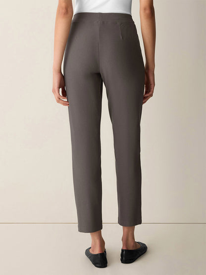 A person stands facing away against a neutral background, wearing the Eileen Fisher Washable Stretch Crepe Slim Pant in Cobblestone—an ankle-length gray style paired with a white top and black flats; the washable fabric ensures easy care.