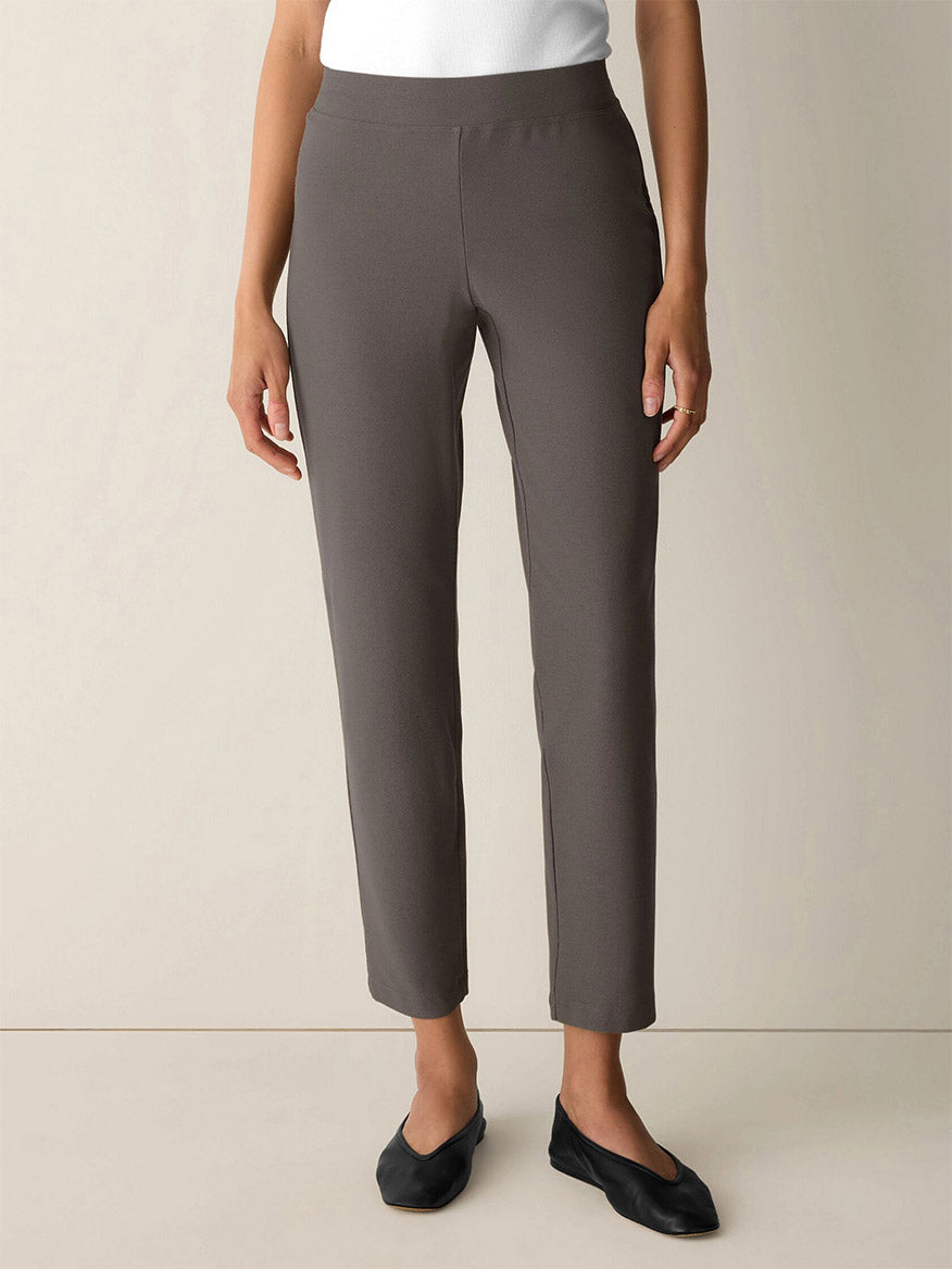 A woman stands against a plain background wearing the Eileen Fisher Washable Stretch Crepe Slim Pant in Cobblestone, paired with a white top and black flats. These pants are slim-fitting and washable for easy care.