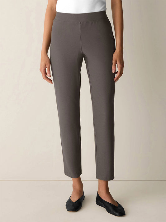 A woman stands against a plain background wearing the Eileen Fisher Washable Stretch Crepe Slim Pant in Cobblestone, paired with a white top and black flats. These pants are slim-fitting and washable for easy care.