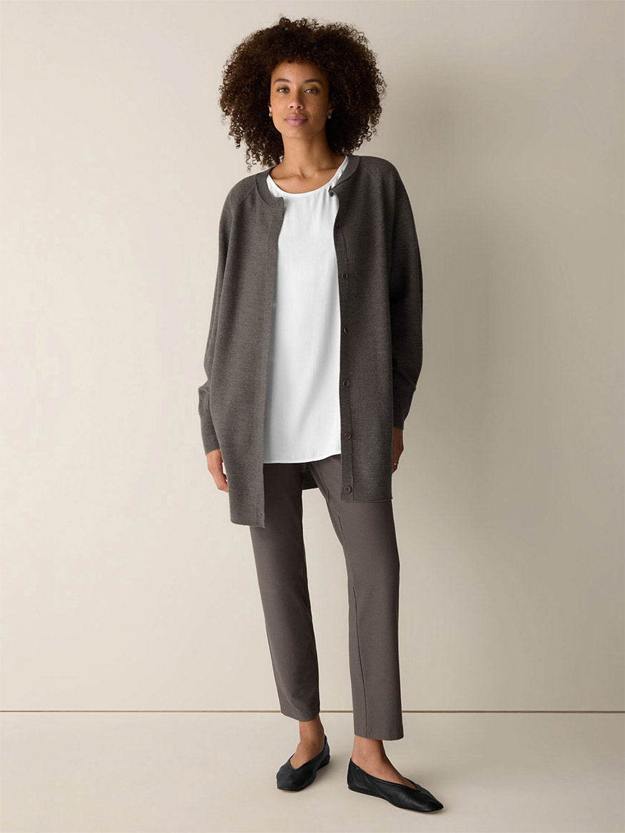 A woman stands against a plain background, wearing a dark gray stretch crepe cardigan, a white blouse, Eileen Fisher Washable Stretch Crepe Slim Pant in Cobblestone, and black flats.