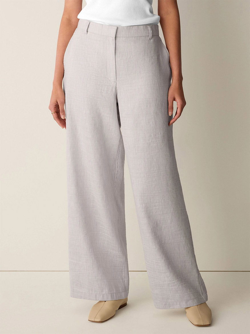 A person wears the Eileen Fisher Striped Linen Sheen Full-Length Trouser in Wheat, paired with a white top, beige shoes, and a striped organic linen jacket with welt pockets and a classic collar against a neutral background.