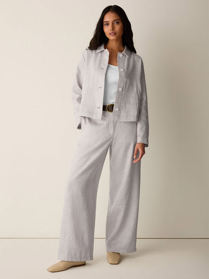 A woman stands against a plain background wearing the Eileen Fisher Striped Linen Sheen Full-Length Trouser in Wheat, paired with an organic linen jacket, white top, black belt, and beige shoes.