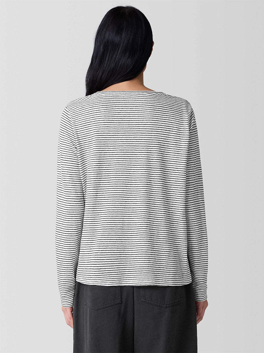 A long-haired person wears an Eileen Fisher Striped Organic Linen Jersey Long-Sleeve Tee in White/Black, paired with dark pants, facing away.